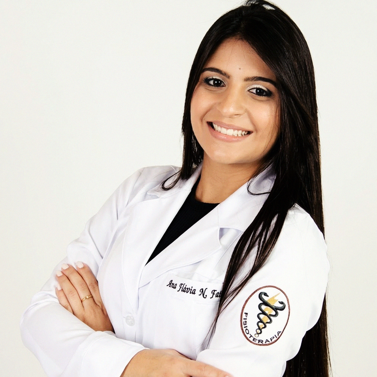 Retrato de meio corpo da Professora Ana Flávia Nunes Faiad. Ela sorri, tem cabelos longos e escuros e está com os braços cruzados. Veste um jaleco branco de fisioterapia com seu nome bordado no peito e o brasão da Fisioterapia com o símbolo da serpente e o raio no braço esquerdo. O fundo é branco e bem iluminado.