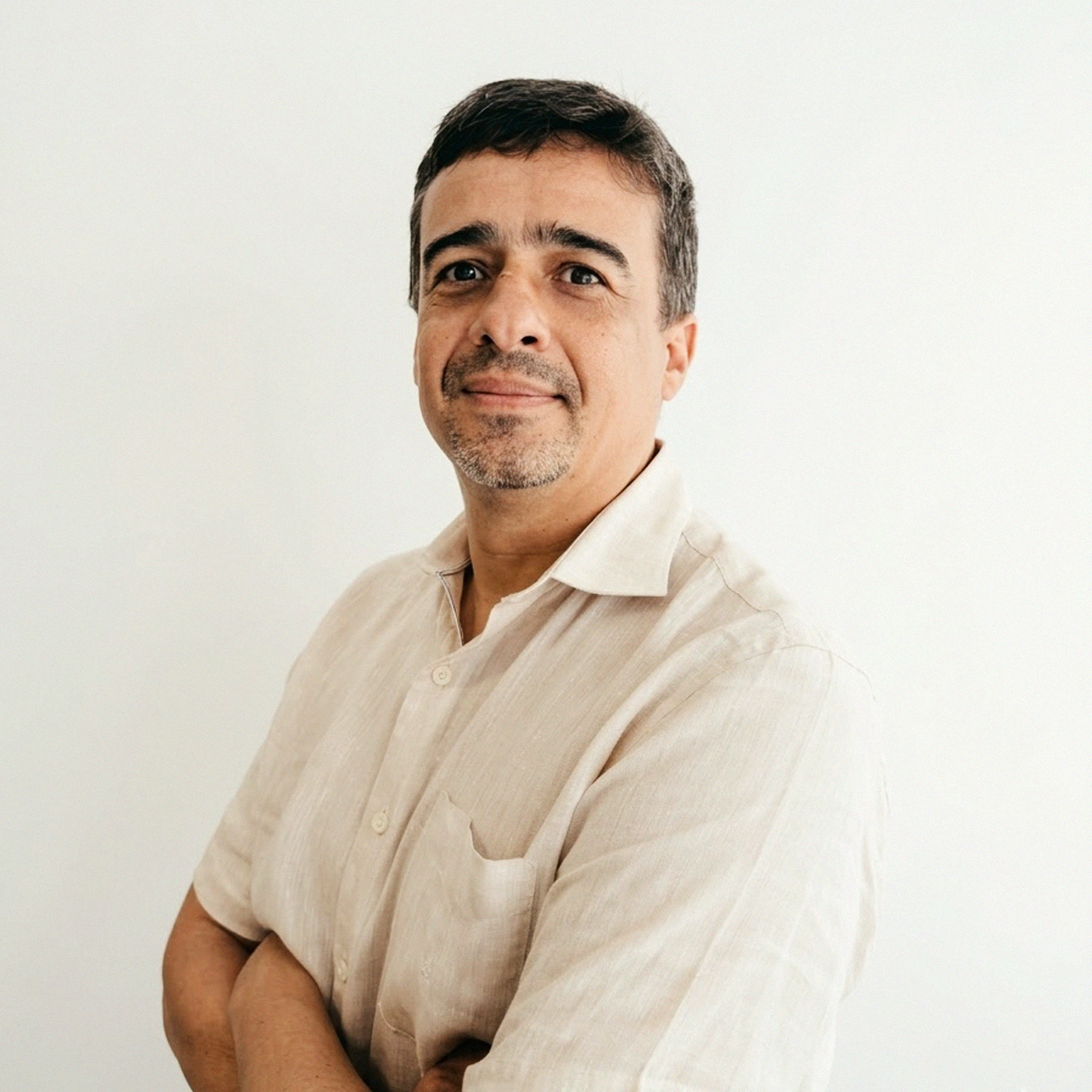 Retrato de perfil do Professor Bruno Porto Pessoa. Ele aparece de braços cruzados, sorrindo levemente e olhando para a câmera. Veste uma camisa de botões em tom bege claro (linho). Homem de cabelos curtos escuros levemente grisalhos e barba por fazer. O fundo é branco, proporcionando um aspecto limpo e profissional.