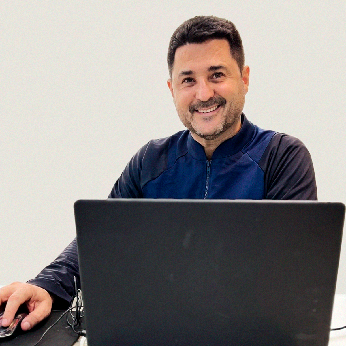 Carlos Henrique Silva Tonázio, homem de pele clara, cabelos escuros curtos e barba aparada, sorrindo para a câmera. Ele está sentado em uma mesa de escritório clara, posicionado atrás de um notebook preto fechado, enquanto segura um mouse com a mão direita. Ele veste uma jaqueta esportiva com tons de azul marinho e preto. A iluminação é clara e uniforme, com um fundo neutro, transmitindo uma imagem profissional e acolhedora.