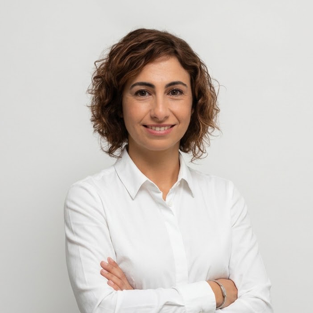 Retrato da professora Cristianne Parizzi Caldeira Brant, com cabelos castanhos curtos e cacheados, vestindo camisa branca de mangas longas. Está sorrindo, com os braços cruzados, diante de um fundo cinza-claro.