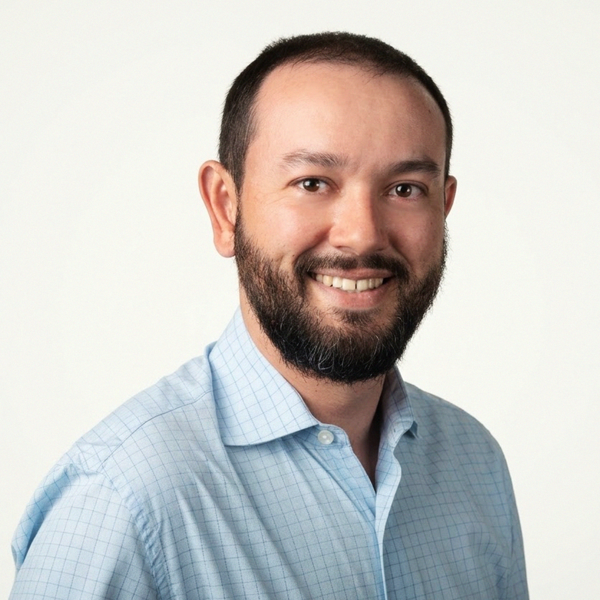 Retrato de close-up do Professor Daniel da Cunha Ribeiro. Ele é um homem de pele clara, com barba e bigode castanhos bem aparados, sorrindo para a foto. Veste uma camisa social de botões em tom azul-claro com estampa de micro-xadrez (grid). O fundo é branco e iluminado, destacando o perfil profissional do docente.
