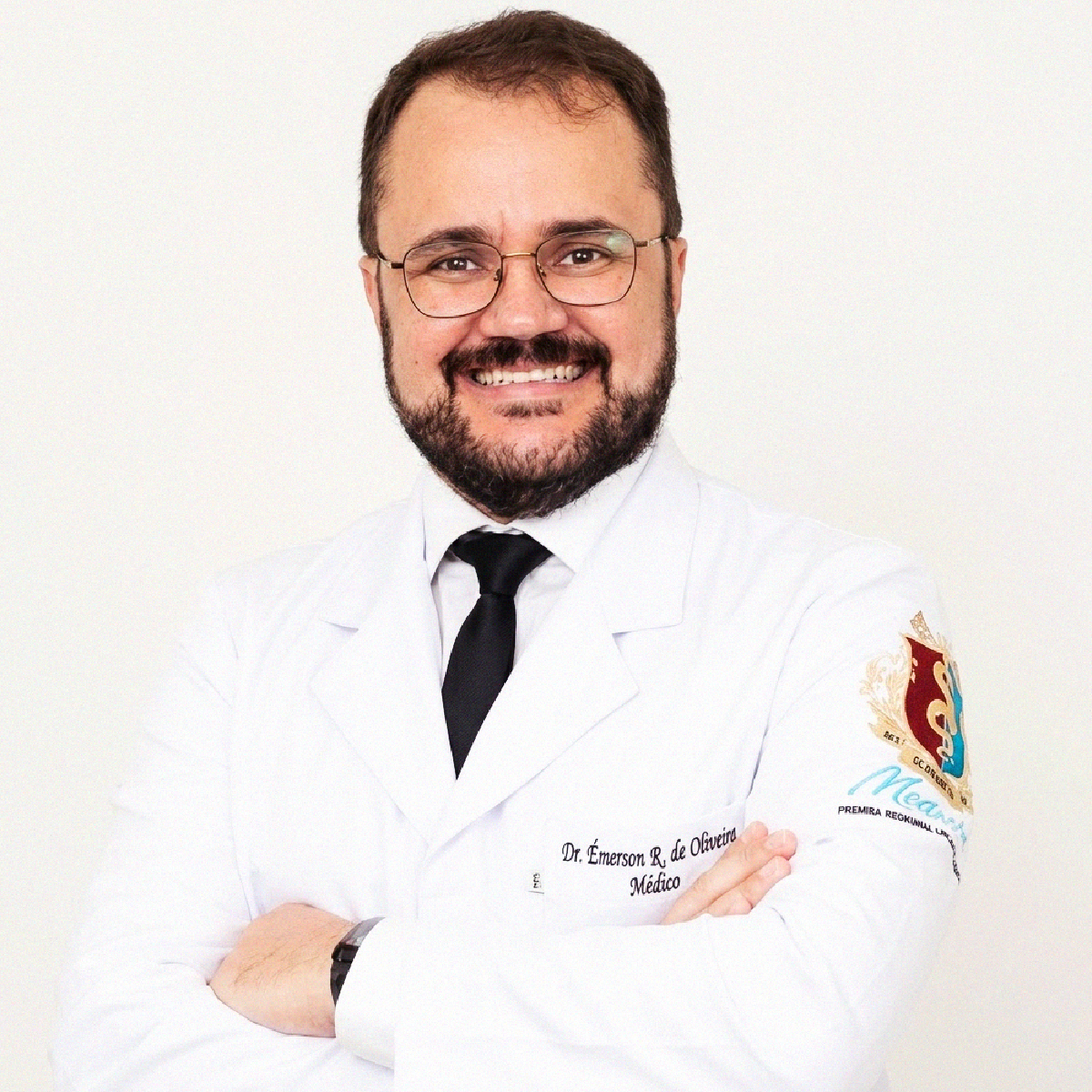 Retrato profissional do Dr. Émerson Fidélis de Oliveira sorrindo, de óculos, jaleco branco e gravata preta, com braços cruzados.