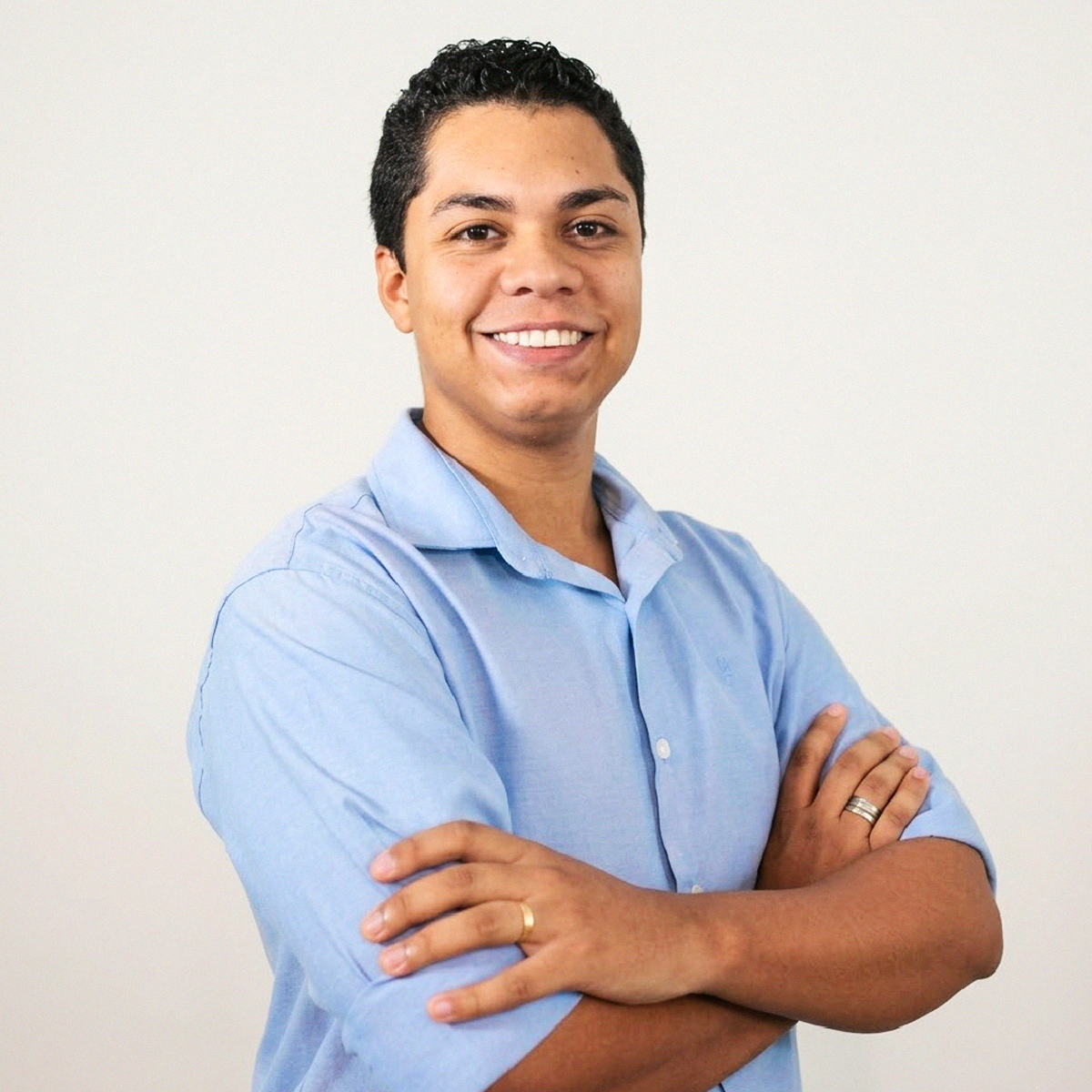 Retrato de close-up do Professor Felipe Paulino do Nascimento, de meio corpo. Ele é um homem de pele parda, com cabelos pretos curtos e encaracolados, sorrindo abertamente para a câmera. Veste uma camisa social de botões em tom azul-claro. Está com os braços cruzados, e nota-se uma aliança de ouro em seu dedo anelar esquerdo e um anel de prata no anelar direito. O fundo é branco e neutro.