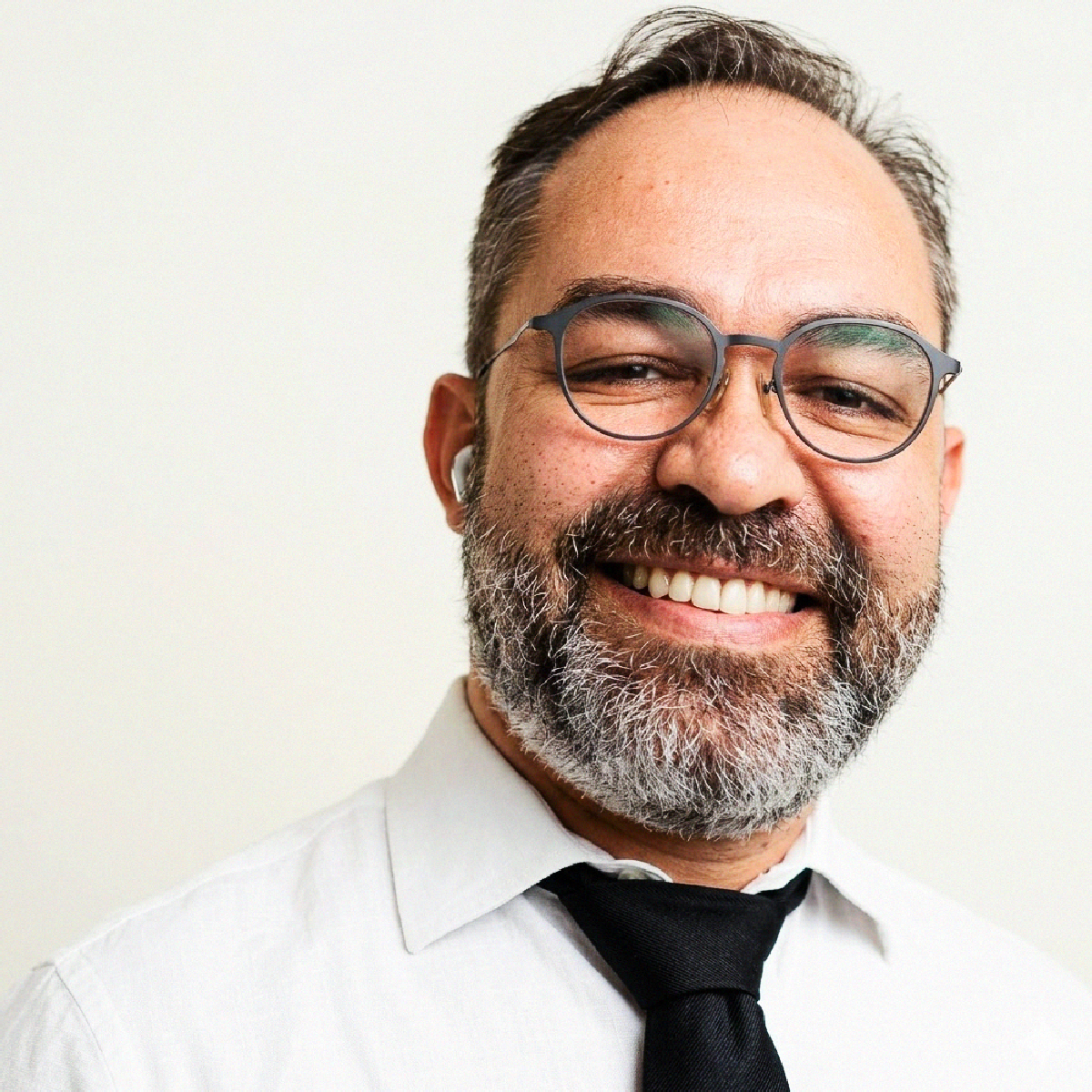 Retrato profissional do Dr. Galeno Hassen Sales sorrindo, de óculos, barba grisalha e fone de ouvido branco no ouvido esquerdo, usando camisa branca e gravata preta.