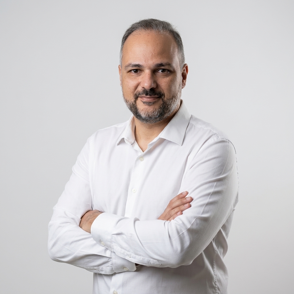 Retrato do professor Geam Karlo de Assis Santana, com cabelos curtos grisalhos, barba curta e expressão serena. Veste camisa social branca e está de braços cruzados diante de fundo cinza-claro.