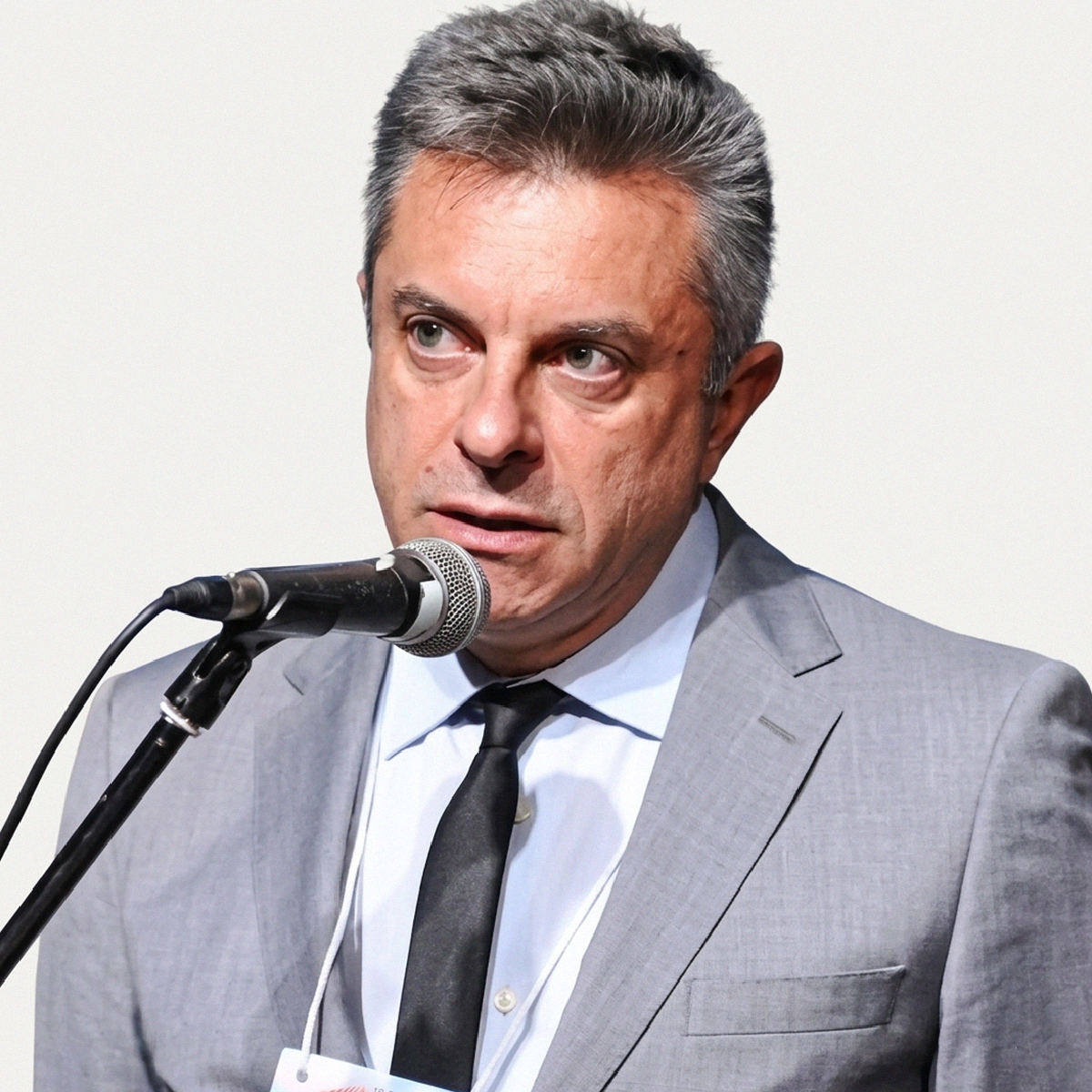 Fotografia de busto do Dr. Jose Milton Cardoso Junior, médico geriatra e professor da FCM-MG, durante uma palestra. Ele veste um terno cinza texturizado, camisa azul clara e gravata preta, posicionado em frente a um microfone preto em um suporte, com fundo branco. Ele está olhando para a direita com expressão séria e atenta, com os lábios ligeiramente abertos. O crachá da instituição é parcialmente visível.