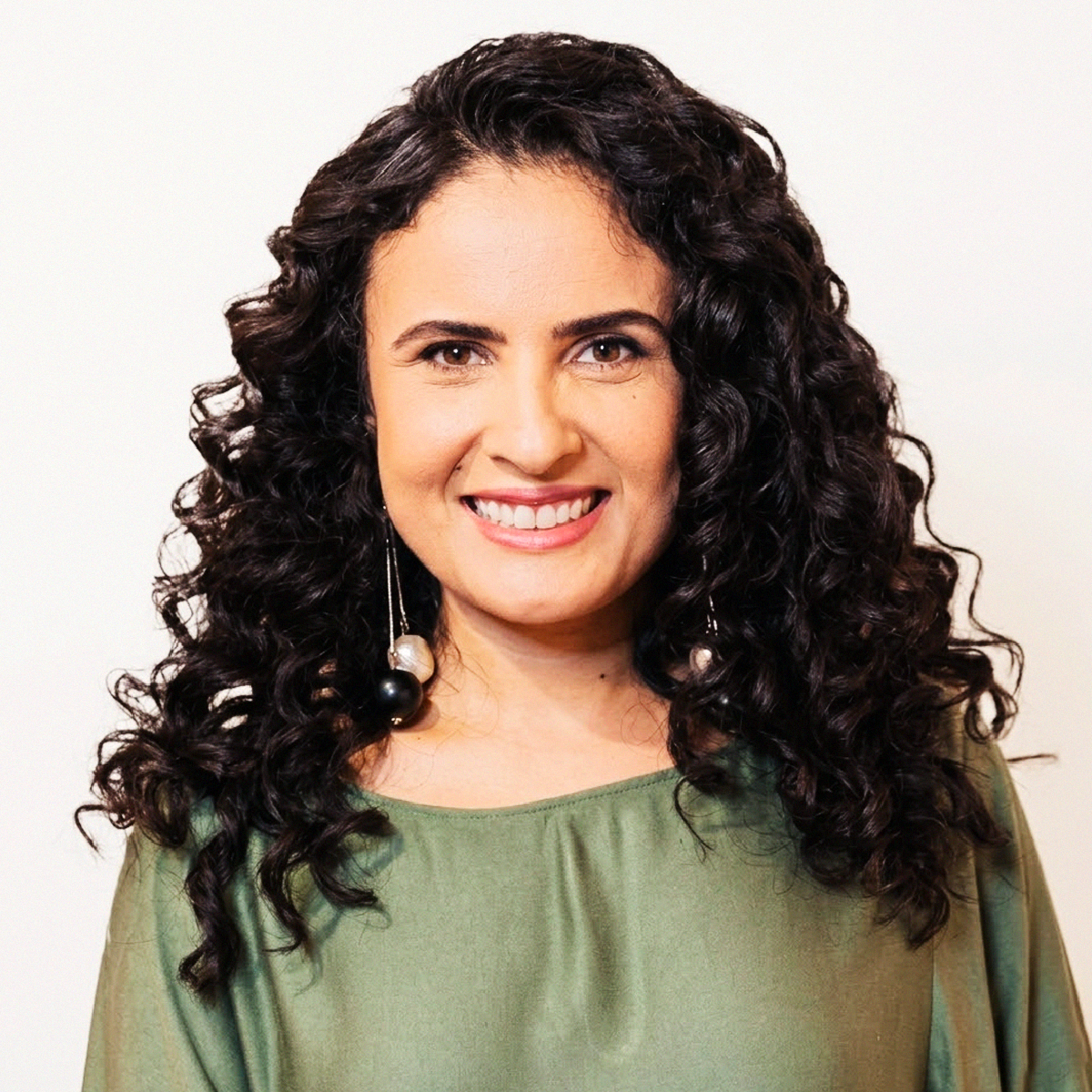 Retrato de rosto da Professora Laís Xavier Vilela. Ela tem a pele clara, olhos castanhos e cabelos pretos, longos e cacheados. Está sorrindo e usa brincos pendentes pretos e brancos em formato de esferas. Veste uma blusa em tom verde-oliva. O fundo é branco e liso.