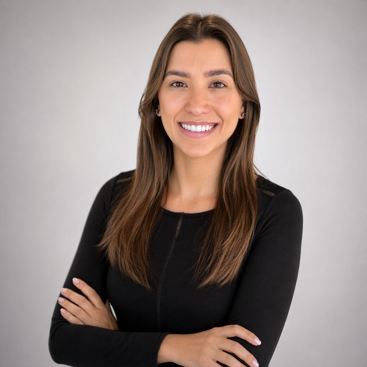 Retrato da professora Larissa Tavares Aguiar, sorrindo para a câmera, com os braços cruzados. Ela tem cabelos longos e castanhos e veste blusa preta de mangas compridas. A fotografia tem fundo claro e neutro.