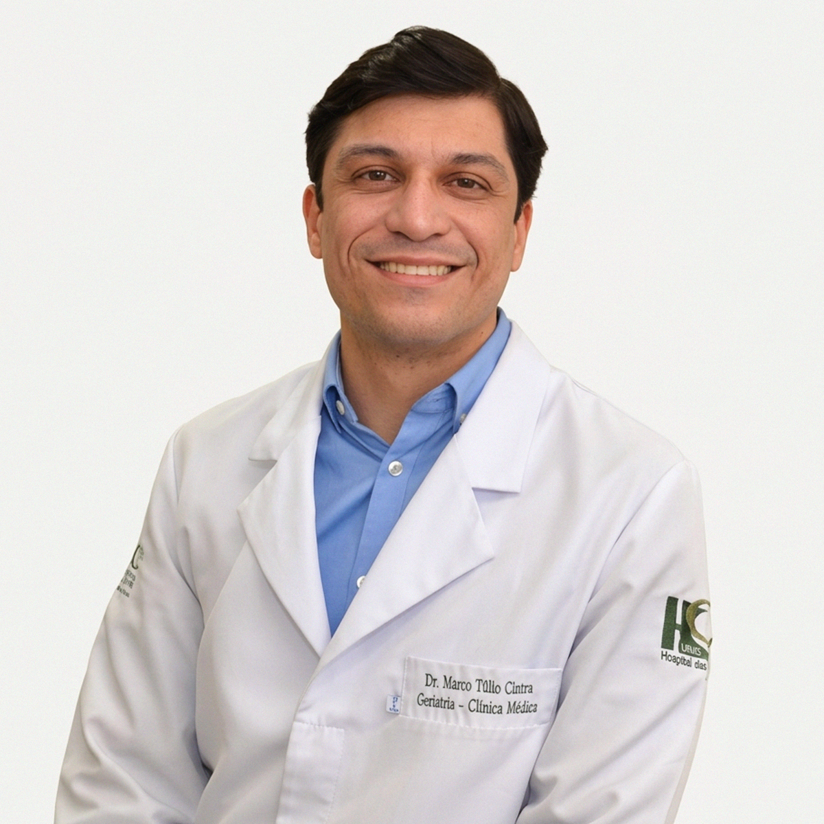 Retrato profissional de meio corpo do Dr. Marco Túlio Gualberto Cintra, médico geriatra e professor. Ele é um homem de pele clara, cabelos pretos curtos e sorri para a foto. Veste um jaleco branco sobre uma camisa social azul clara. No bolso do jaleco, há um bordado com seu nome e as especialidades "Geriatria - Clínica Médica". Na manga esquerda, vê-se o logotipo do Hospital das Clínicas da UFMG. O fundo é branco e bem iluminado.