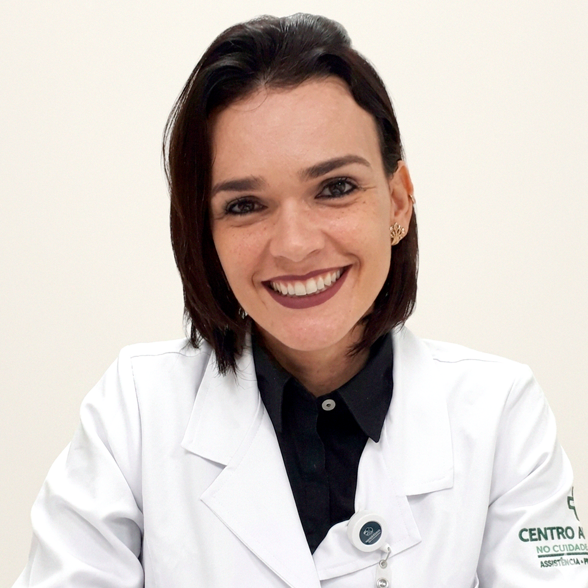 Fotografia de perfil de Maria Clara Salomão e Silva Guimarães, enfermeira e docente da FCM-MG. Ela é uma mulher de pele clara, cabelos castanhos curtos, sorridente, vestindo jaleco branco com o emblema do "Centro de Cuidados" no braço e camisa preta por baixo. O fundo é neutro e claro.