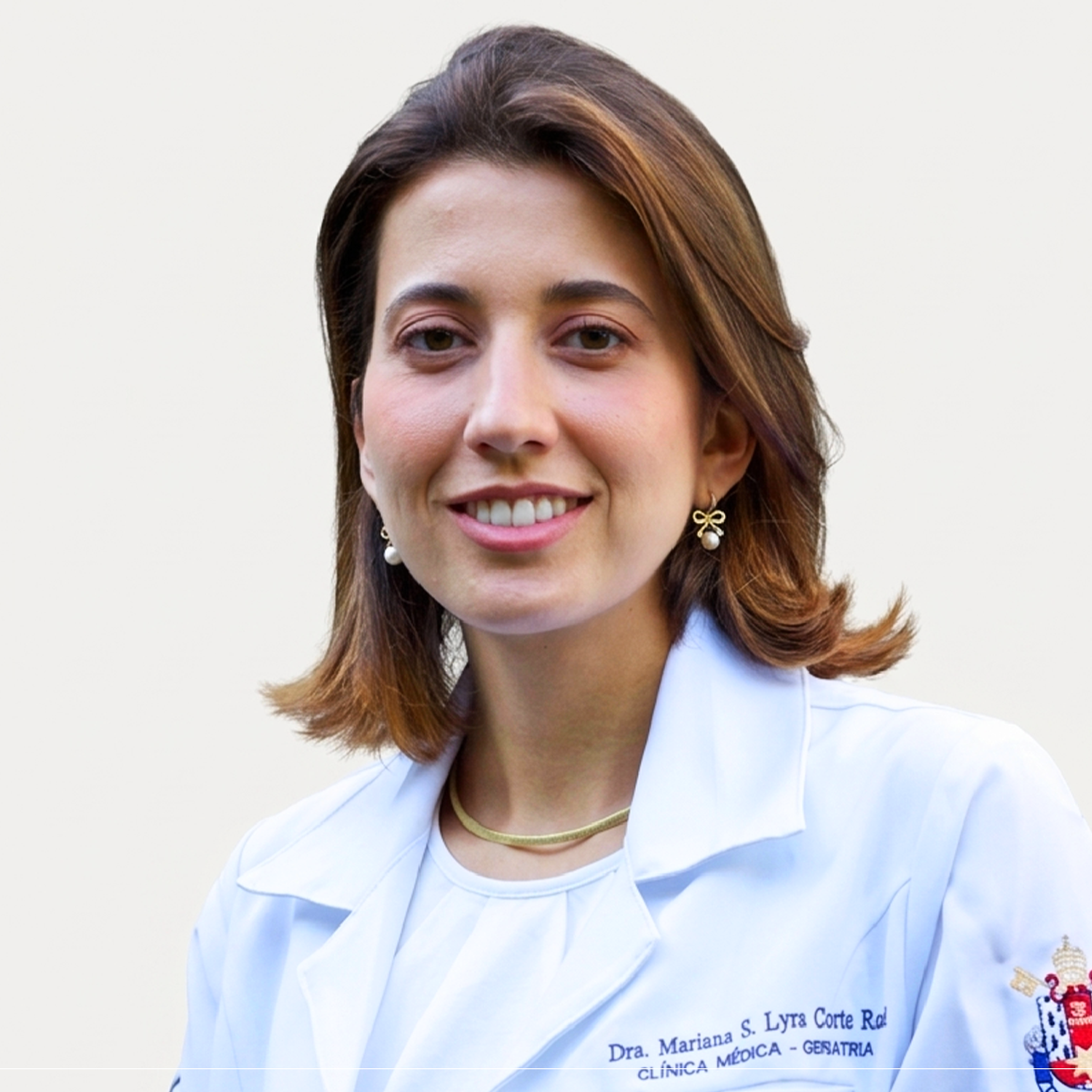 Retrato profissional de busto da Dra. Mariana Santos Lyra Corte Real, médica geriatra e professora. Ela tem pele clara, cabelos castanhos na altura dos ombros e sorri para a foto. Veste um jaleco branco com gola estruturada; no lado direito do peito, lê-se o bordado azul "Dra. Mariana S. Lyra Corte Real - CLÍNICA MÉDICA - GERIATRIA". Na manga esquerda, há um brasão institucional colorido. Ela usa brincos pequenos em formato de laço dourado com uma pérola pendente e um colar de malha dourada fina. O fundo é neutro e claro.