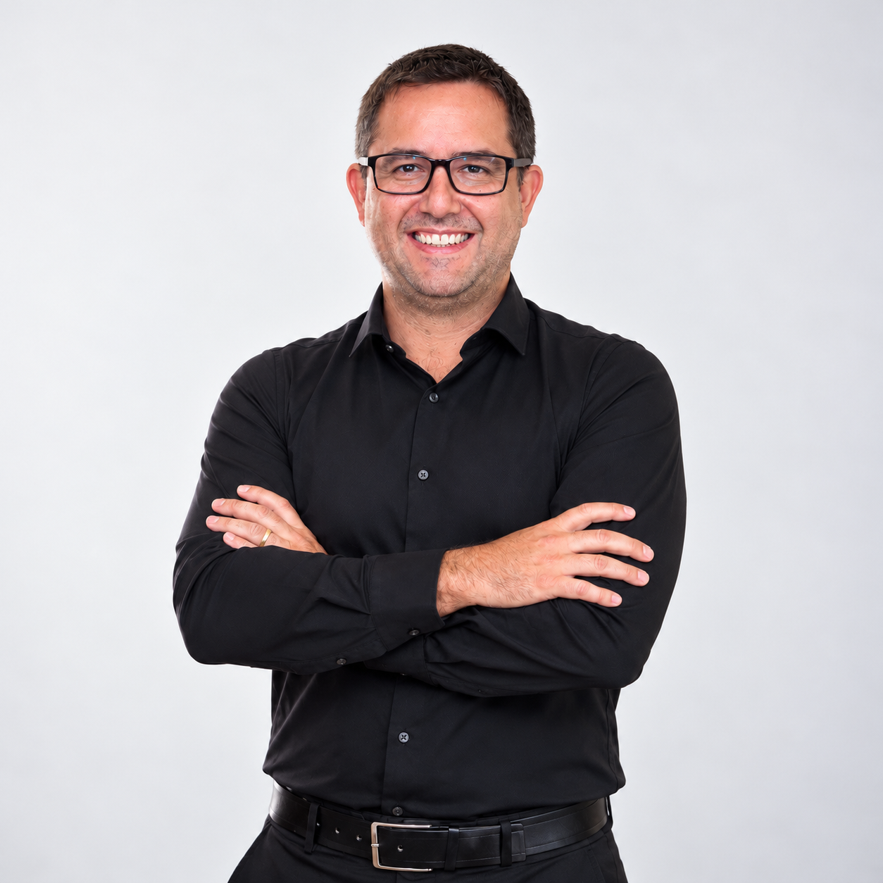 Retrato do professor Rafael Duarte Silva, sorrindo, com cabelos castanhos curtos, óculos de armação preta e camisa social preta, de braços cruzados, diante de fundo claro.
