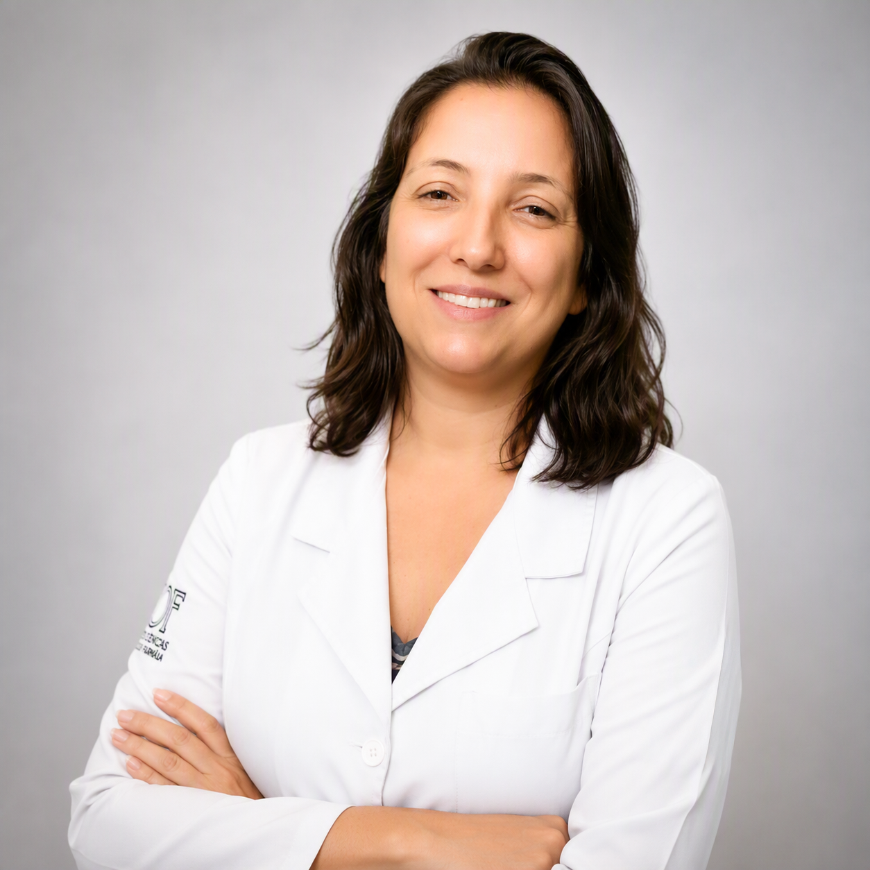 Retrato da professora Raquel de Carvalho Lana Campelo, sorrindo, com os braços cruzados. Ela tem cabelos escuros na altura dos ombros e veste jaleco branco, diante de fundo claro e neutro.