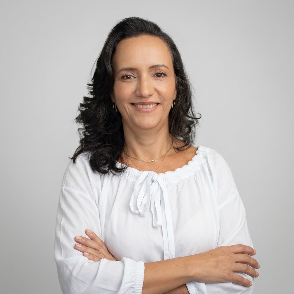 Retrato da professora Silvia Elizate Monteiro, com cabelos escuros, longos e ondulados, vestindo blusa branca de mangas compridas, com amarração na gola. Aparece sorrindo, com os braços cruzados, diante de um fundo cinza-claro.