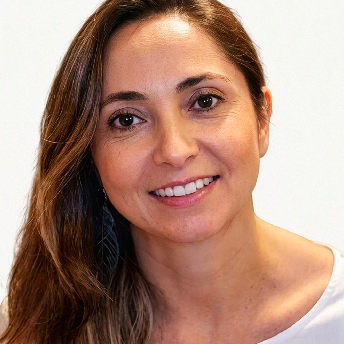 Retrato de close-up da Professora Sonaly Cristine Leal. Ela possui pele clara, olhos castanhos e cabelos castanhos longos e levemente ondulados. Está sorrindo suavemente para a câmera. Usa um brinco pendente escuro em formato de folha ou pena na orelha direita. Veste uma blusa branca de decote canoa. O fundo é claro e neutro.