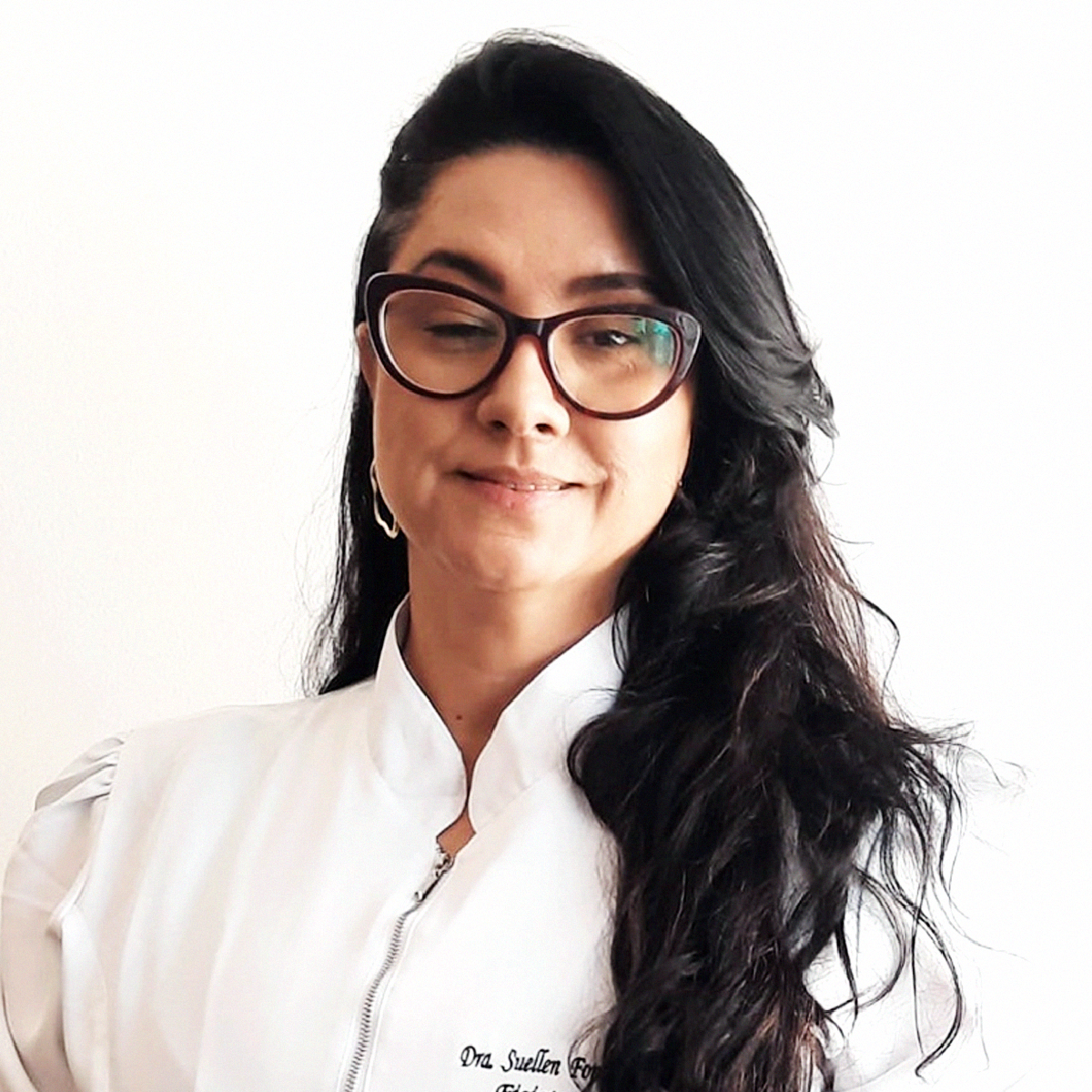 Retrato de rosto da Professora Suellen Fonsêca Santos. Ela possui cabelos pretos longos e ondulados, pele clara e usa óculos de grau com armação escura em formato "gatinho". Veste um jaleco branco com gola padre e detalhe de zíper frontal, onde se lê o bordado "Dra. Suellen" no lado esquerdo do peito. O fundo é branco e neutro.