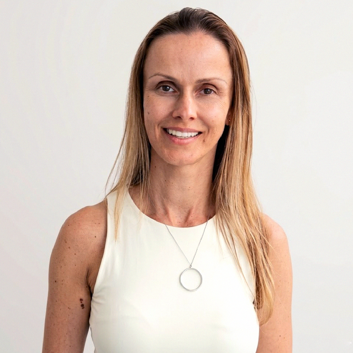 Retrato de close-up da Professora Tatiana Péret Barbosa. Ela possui pele clara, olhos castanhos e cabelos loiros lisos na altura dos ombros. Está sorrindo para a câmera e veste uma blusa off-white de decote careca (gola redonda). Usa um colar fino de metal prateado com um pingente circular liso. O fundo é branco e liso.