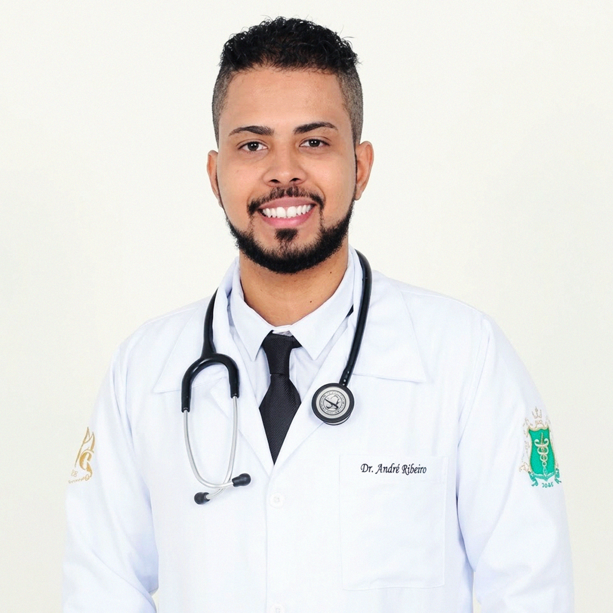 Retrato de plano médio do Professor André Ribeiro de Paula. Homem jovem, com barba aparada e sorriso expressivo, veste jaleco branco com estetoscópio preto sobre os ombros. No lado esquerdo do peito, lê-se o bordado "Dr. André Ribeiro". O jaleco possui brasões coloridos bordados em ambas as mangas, sendo o da esquerda um símbolo verde de saúde. Fundo branco limpo.