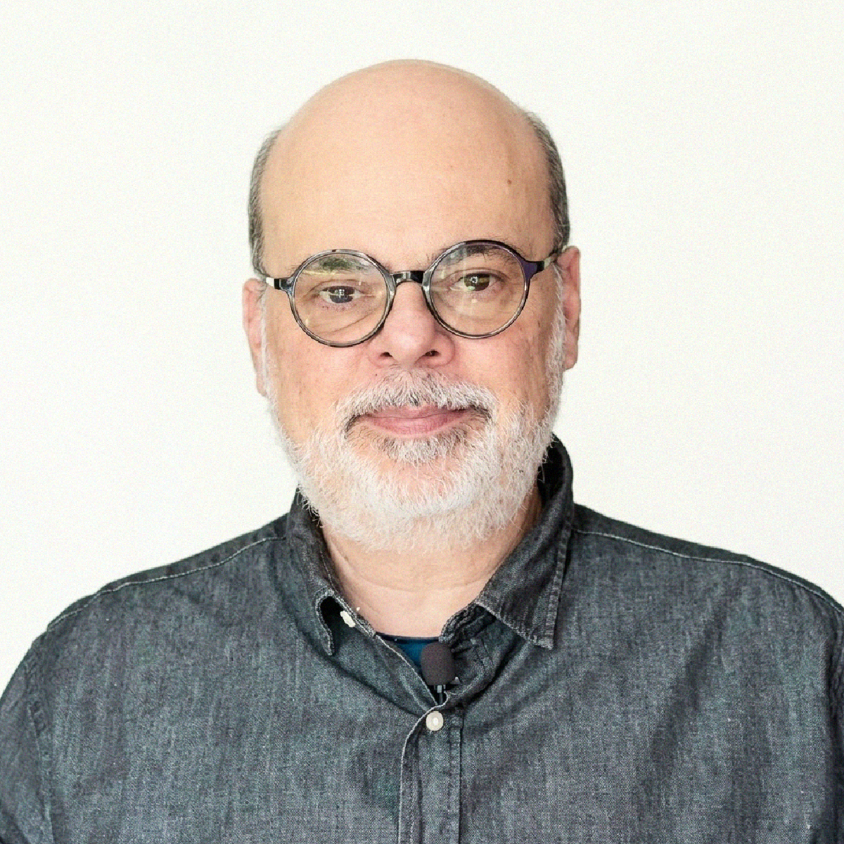 Retrato profissional de meio corpo do Prof. Dr. Antônio Carlos de Castro Toledo Júnior, médico infectologista e professor da FCM-MG. Ele é um homem calvo, com barba branca curta, pele clara e usa óculos redondos com armação escura metálica. Veste uma camisa de botão cinza texturizada. Um pequeno microfone lapela preto está preso à camisa. Ele olha diretamente para a câmera com um sorriso leve e amigável. O fundo é branco liso e bem iluminado.