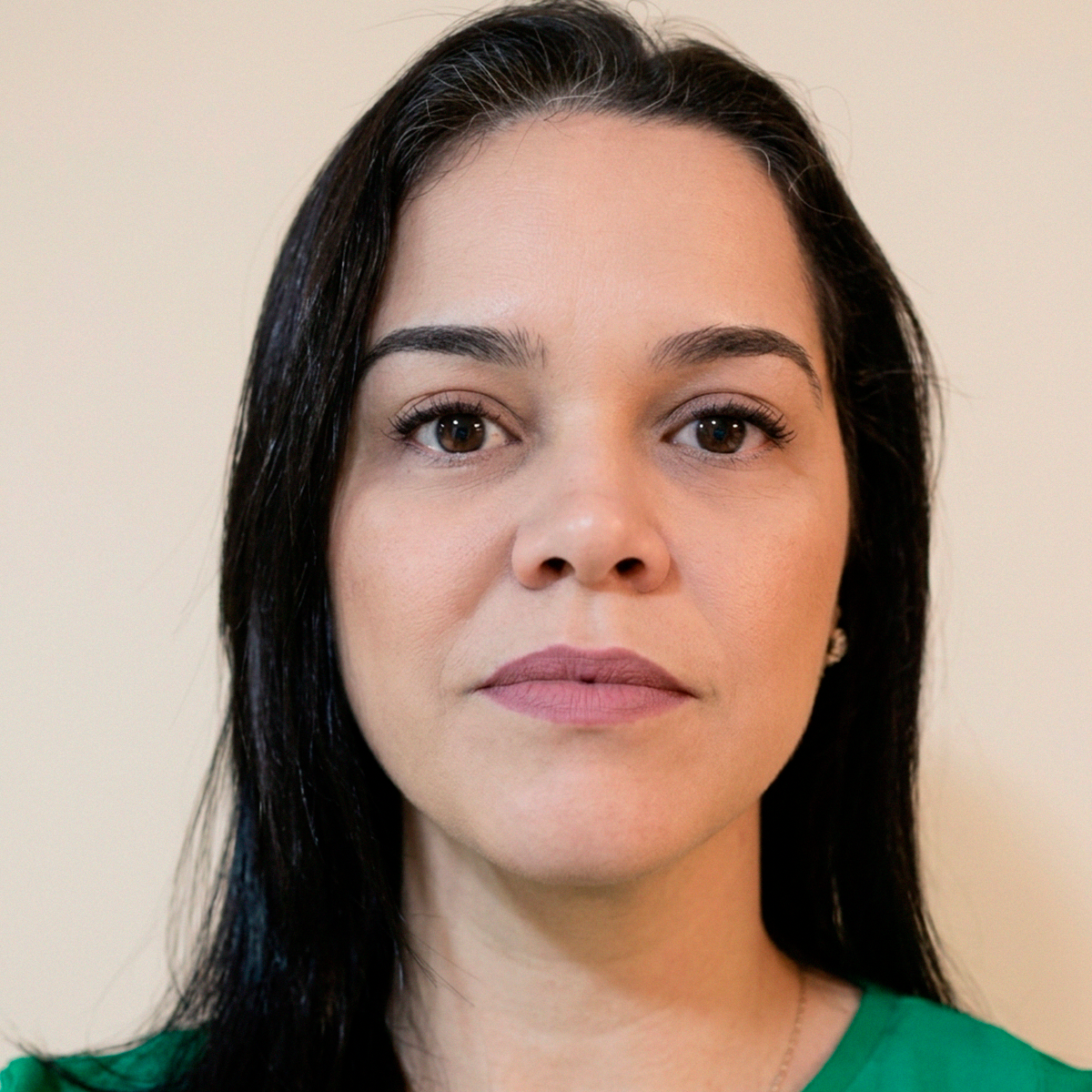Shirlei Barbosa Dias, mulher de pele clara, com longos cabelos escuros e levemente grisalhos na raiz, olhando diretamente para a câmera. Ela veste uma blusa verde vibrante e usa um pequeno brinco delicado. A iluminação é suave e o fundo é neutro, em um tom de bege claro, transmitindo uma imagem profissional e serena.