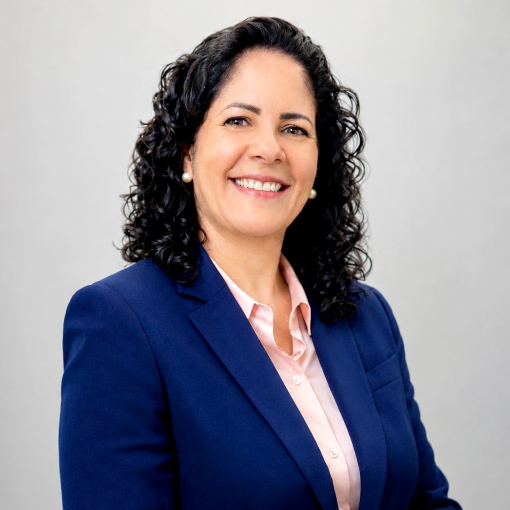 Foto profissional de Meire Rose de Oliveira Loureiro Cassini, uma mulher sorridente com cabelos pretos cacheados na altura dos ombros. Ela veste um blazer azul-marinho sobre uma camisa social rosa clara e usa brincos de pérola discretos. A imagem é um retrato de meio corpo (headshot) com um fundo cinza claro e neutro.