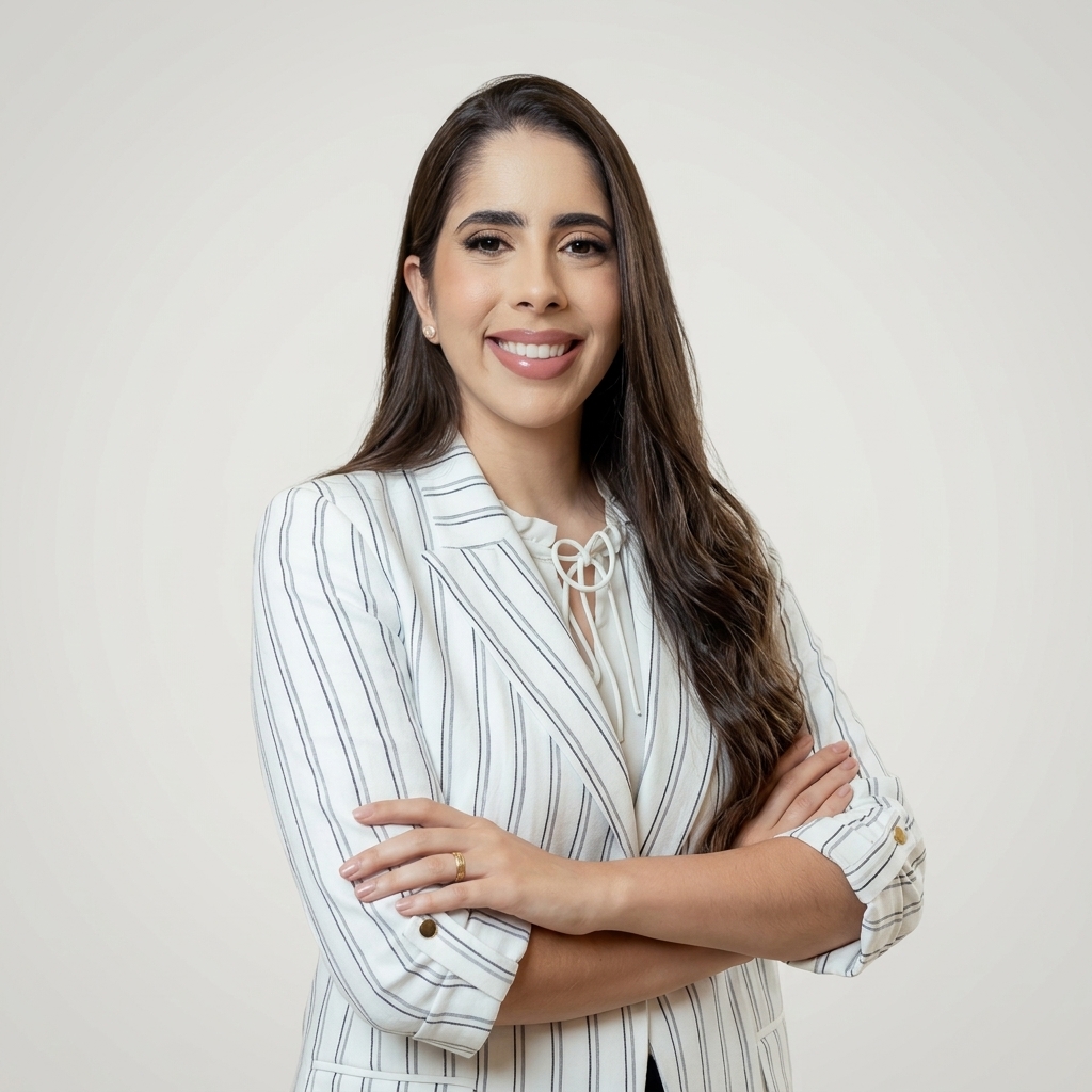 Retrato de Bruna Pinto Gomes, médica ginecologista e obstetra, com cabelos castanhos longos, blazer branco listrado e braços cruzados, sorrindo diante de fundo claro.