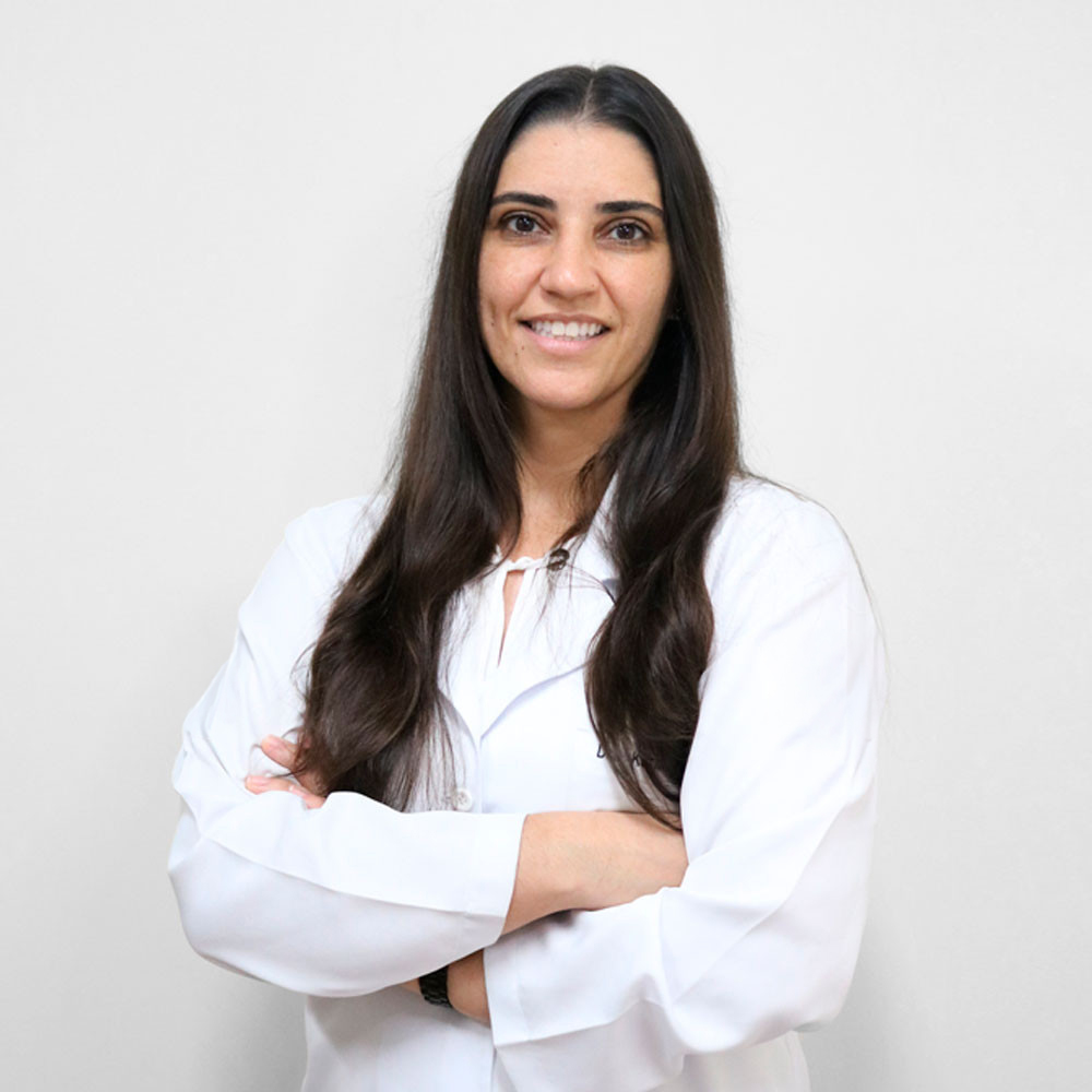 Retrato da médica Karla de Carvalho Schettino, com cabelos castanhos escuros, longos e lisos, vestindo jaleco branco, com os braços cruzados e expressão sorridente diante de fundo claro.